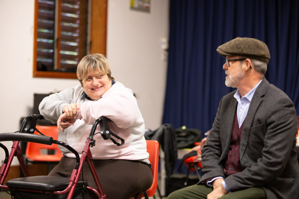 a woman is sittind down, leaning on a walking aid and smiling. a man in a flat cap sits next to her
