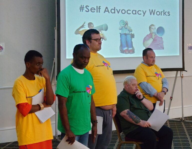 a group of people in bright T shirts listen while standing in front of a screen that says # self advocacy works