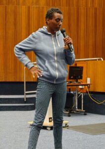 A woman in a grey hooded top speaks into a microphone with one hand on her hip
