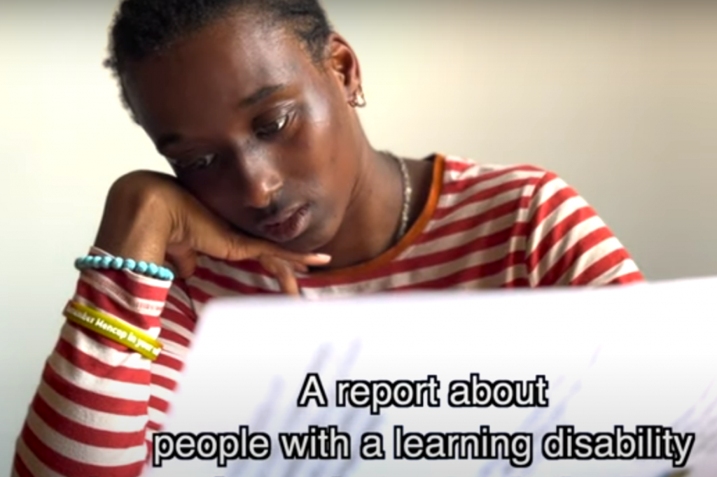 A young black woman reads a document. The caption on the screen says 'A report about people with a learning disability'.