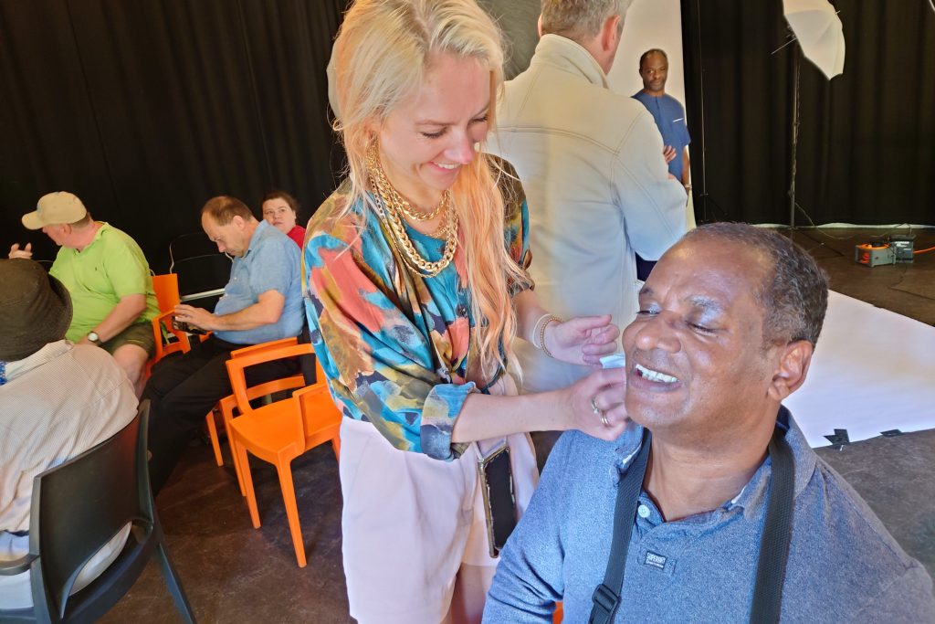 A woman is standing applying make up to a man's face. He is sitting in a chair beside her.