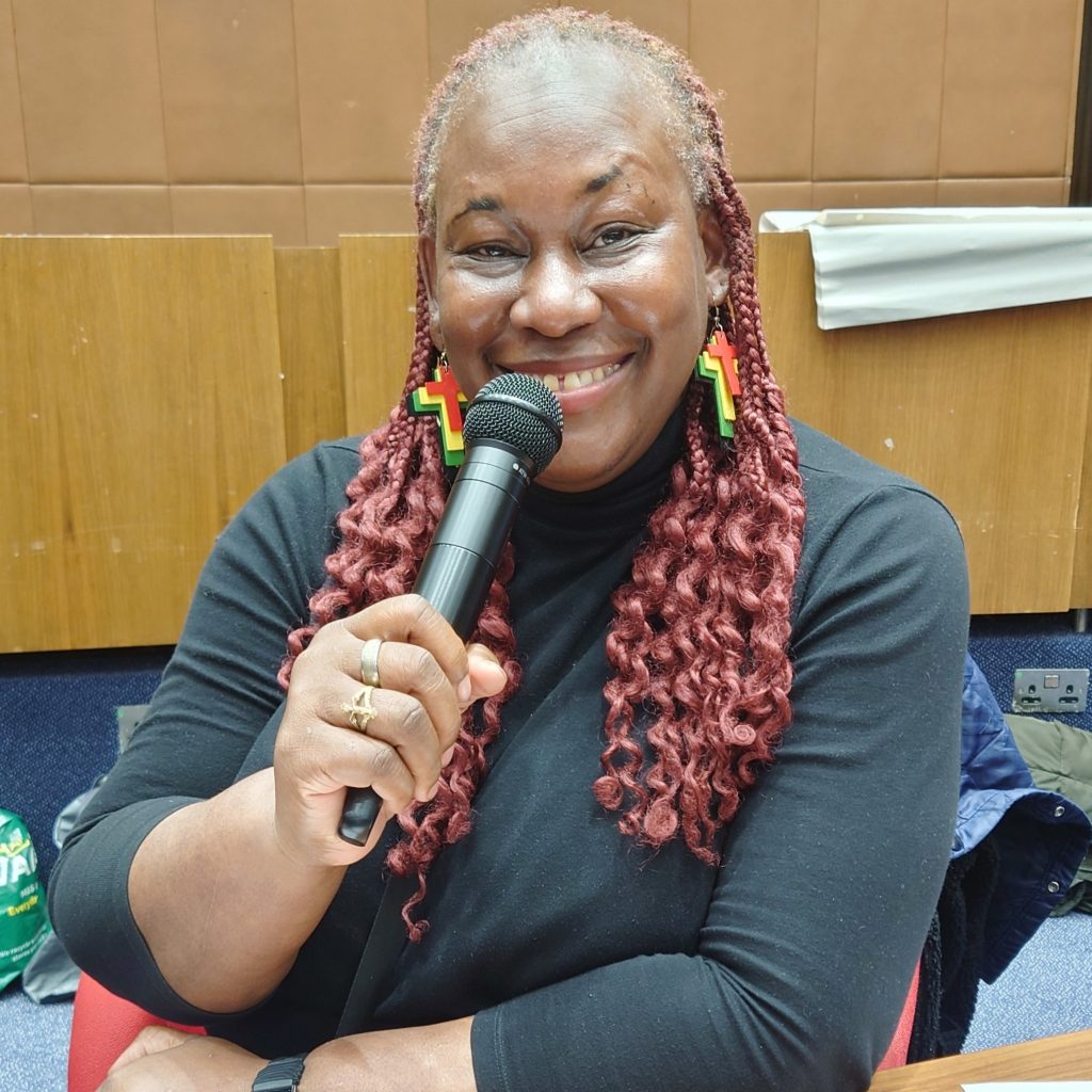 A black woman is holding a microphone. She is smiling and looking towards the camera.